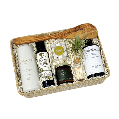 housewarming gift that includes an olivewood spatula, multisurface spray, hand soap, olive oil, gummy candies, fir + clove candle, matches, a striped tea towel, small air plant in a woven gift basket