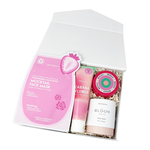 A curated beauty gift set in a white box, featuring a pink strawberry mocktail face mask, Mersea cabana flower lotion and scented candle and a rose scented bath truffle.
