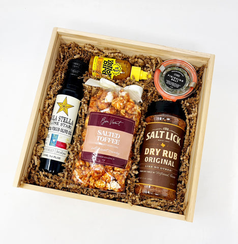 Gift basket with various items including a bottle of olive oil, salted toffee popcorn, finishing salt, hot sauce and a local BBQ Dry rub in a wooden box.