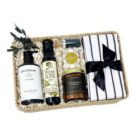 curated housewarming gift basket with small batch products made in texas like hand soap, texas hill country olive oil, a candle, matches, celebratory candy and a striped microfiber tea towel in a keepsake woven seagrass reed gift box.